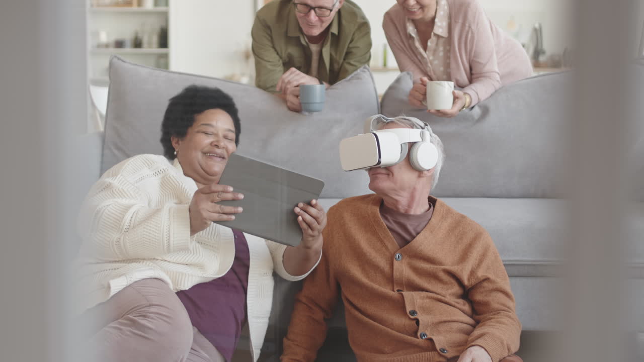 Medium shot of happy diverse senior friends hanging around couch at home, man using VR headset, African woman having tablet computer, grey-haired couple holding cups looking at them