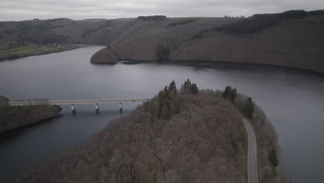 Drohnenaufnahme-In-Luxemburg-In-Der-Nähe-Des-Flusses-Lac-De-La-Haute-Sure-Im-Naturpark-Öewersauer-An-Einem-Bewölkten-Tag-Mit-Auf-Der-Straße-Fahrenden-Autos-In-Der-Nähe-Des-Waldes-Und-Der-Bäume-Und-Einer-Brücke-Im-Hintergrund