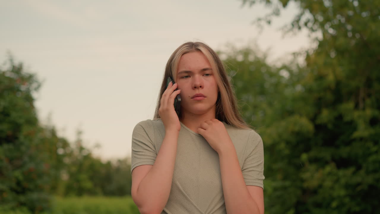 joven con camiseta gris caminando por un camino rodeado de exuberante vegetación, involucrada en una conversación telefónica con expresión reflexiva y una mano tocando su cuello