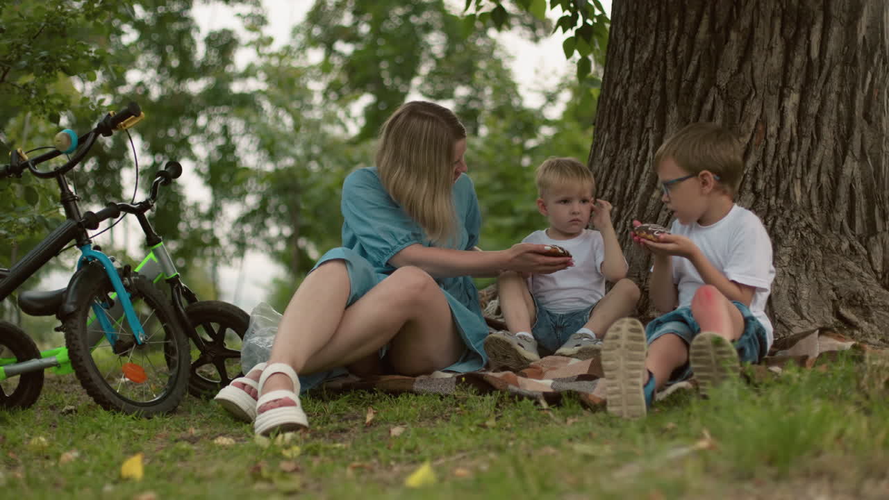 A mother and her two children sit on the ground under a tree, the mother offers a snack to her younger child, who refuses it, pointing towards the older sibling s snack