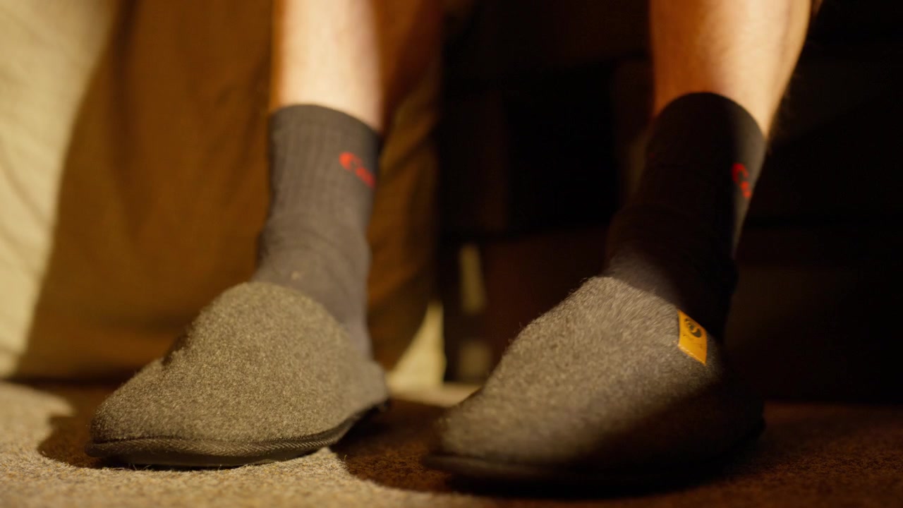 Close-up, persoon met sokken aan stapt uit bed om voeten in warme pantoffels te schuiven op de vloer in een hotel of thuis.