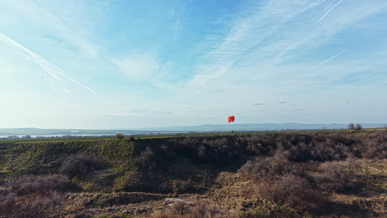 Un parapendio vola sopra colline verdi in una giornata limpida in una zona rurale con cielo blu e nuvole bianche in lontananza.