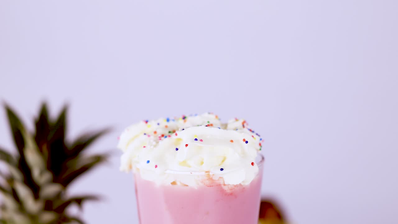 A hand gently places a cherry atop whipped cream and sprinkles on a pink milkshake