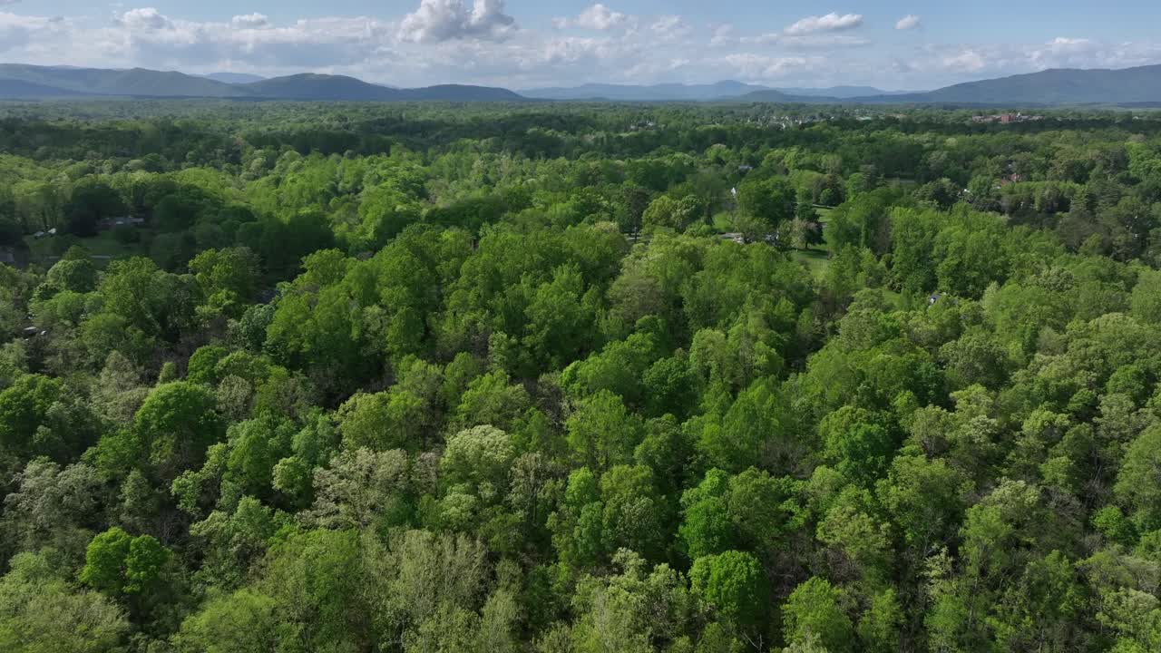 Drone flight over green landscape with forest trees and houses. Mountain range and Clouds at sky. Spring day in United States. Residential area with homes between woodland.