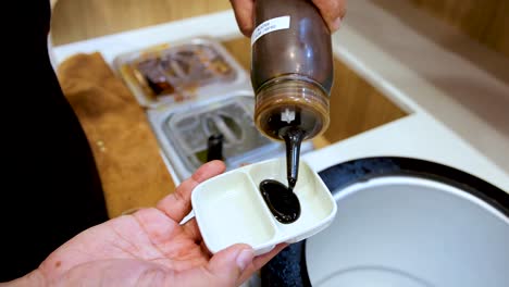 a person pours a thick, dark sauce from a squeeze bottle into a white ceramic dipping dish in a brightly lit kitchen setting