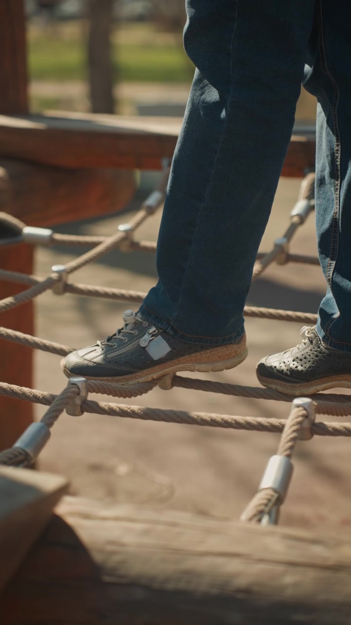Close up of child carefully walking across outdoor rope bridge balancing on each step. focus remains solely on lower legs and feet in motion as they navigate thick ropes secured with metal