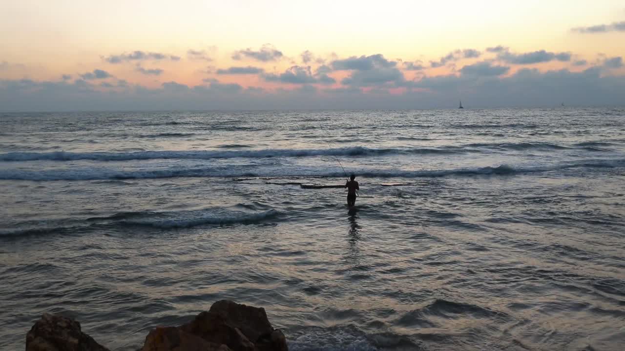 A fisherman walks in the sea, on the beach of Tel Aviv.The beach at sunset, waves breaking on the beachship sails deep into the sea, orange-colored sunset,Tel Aviv beach