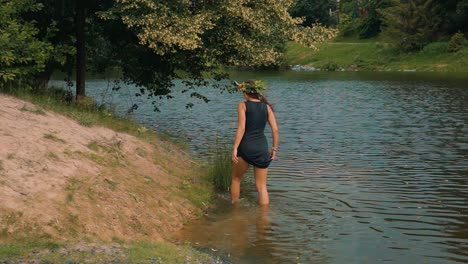 slow motion of attractive barefoot female in dress with flower wreath in nature walking in riverwater and riverbank under tree