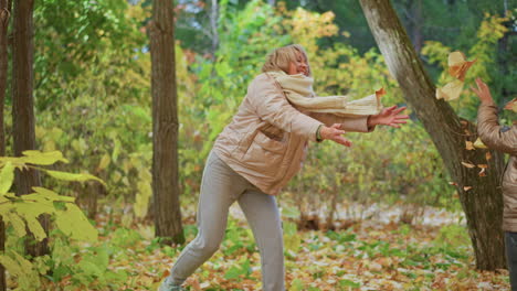 laughing mother stands in forest filled with yellow leaves while girl playfully tosses dry foliage toward her, both dressed warmly enjoying lighthearted moment on crisp bright autumn day outdoors
