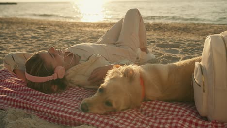 una chica rubia con ropa ligera escucha música con auriculares y se acuesta en una alfombra con su perro en una playa soleada