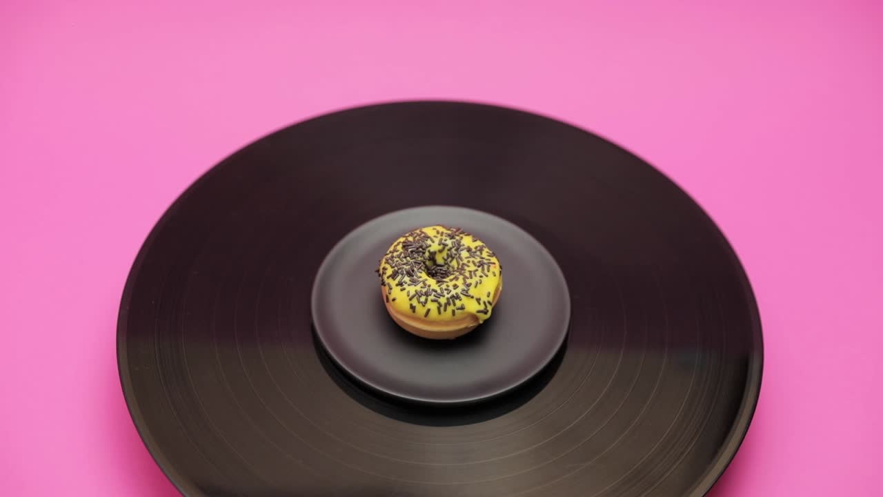 An American donut topped with yellow chocolate with black choco crumbs turns in a circle on a black turntable. The shot from above
