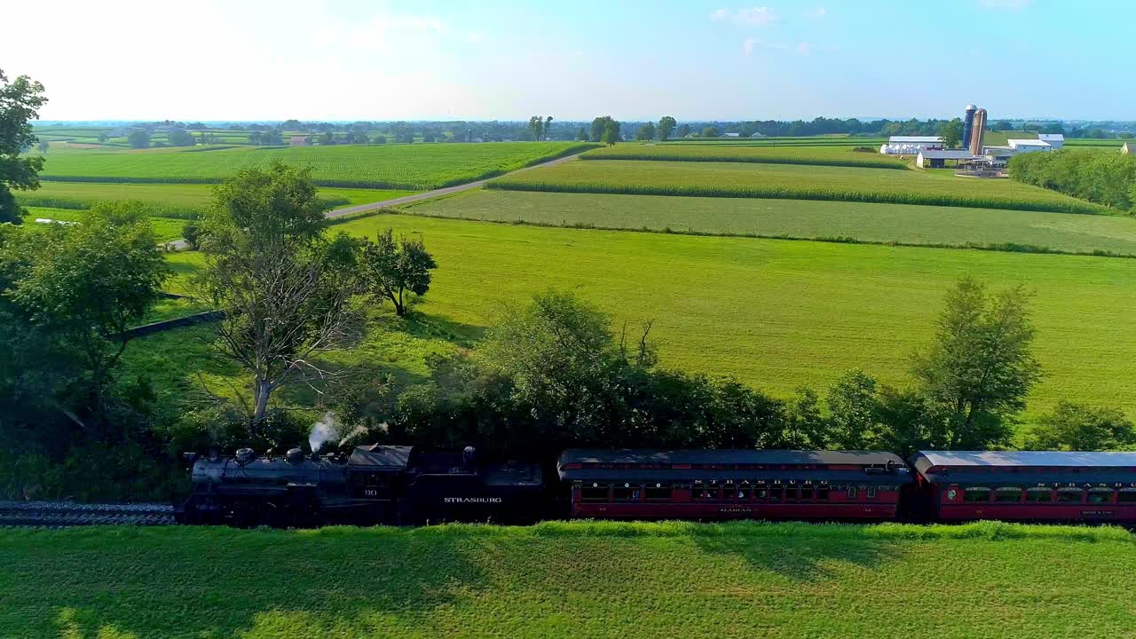 A historic steam train glides along tracks surrounded by lush green fields and trees, showcasing the beauty of rural America in the afternoon light.