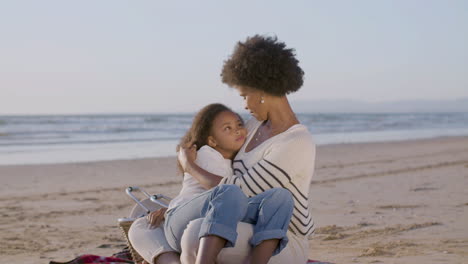 happy woman holding her cute daughter lying on her knees, stroking her curly hair and talking together during a picnic on the beach