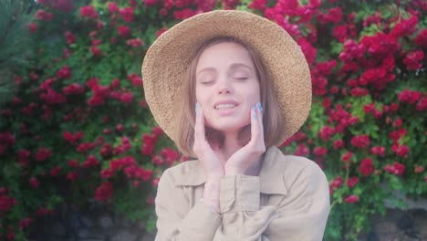 elegante mujer joven rubia en un sombrero de paja en el fondo de un arbusto de rosas en flor