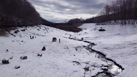 Picturesque-winter-flight-above-people-playing-in-snow-with-dogs-and-kids-by-river's-edge-toward-reflective-lake-water-and-bridge-in-countryside,-overhead-aerial-approach