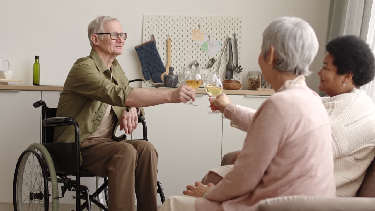Medium long of happy elderly handicapped Caucasian man in wheelchair drinking white wine, clinking glasses and talking to his friends at home
