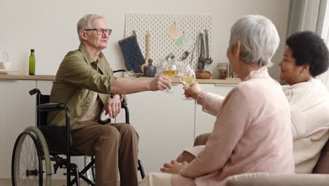 medium long of happy elderly handicapped caucasian man in wheelchair drinking white wine, clinking glasses and talking to his friends at home
