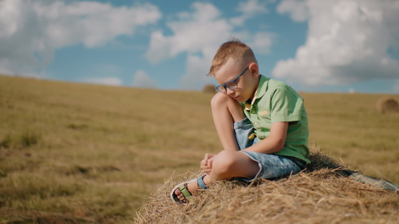 маленький мальчик сидит на пучке сена с наклоненной головой, потерянный в мыслях, окруженный огромным открытым полем под ярким голубым небом, захватывая тихий, мирный момент в сельской местности