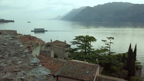 Atemberaubender-Sonnenuntergang-über-Dem-Gardasee-Von-Der-Burg-Von-Malcesine,-Mit-Blick-Auf-Die-Stadtlandschaft,-Zypressen,-Berge-Und-Ruhiges-Wasser,-Vorbeifahrende-Boote-Und-Fähren-Tragen-Zur-Ruhe-Bei