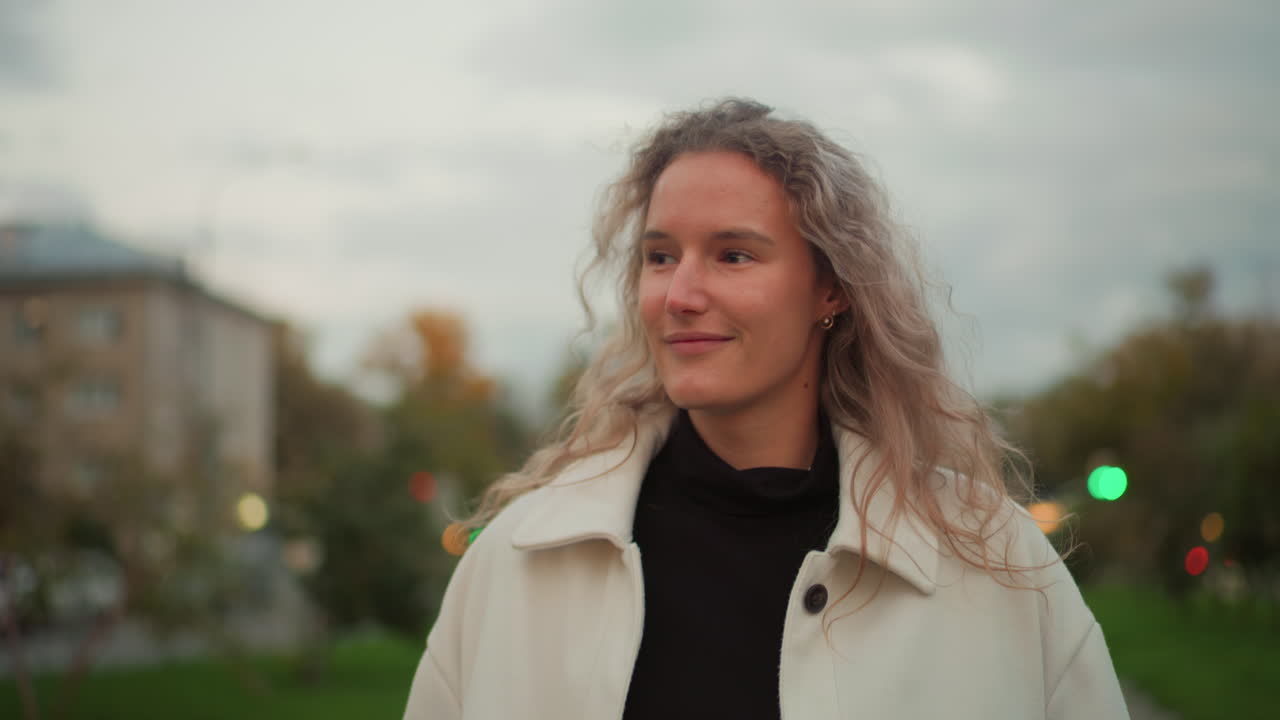 Elegant lady glancing to side with charming smile while holding white umbrella, preparing to twirl it, wearing light coat, with soft background of trees, buildings, and evening lights