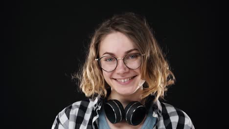 retrato en primer plano de una joven europea sonriendo humildemente a la cámara sobre un fondo negro. sonriendo alegremente. con gafas y auriculares en el cuello