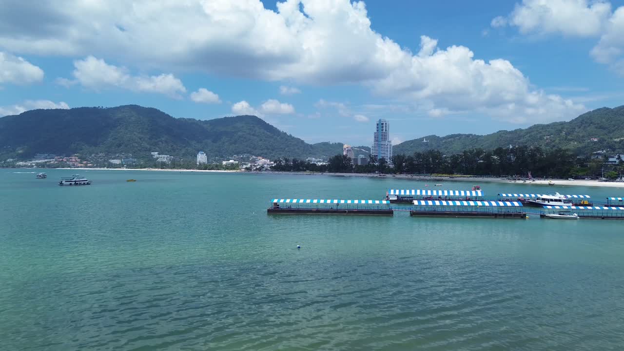 Drone flies up behind a rock, unveiling the stunning Patong Beach with turquoise waters and swimmers enjoying the sunny coastline of Phuket, Thailand.