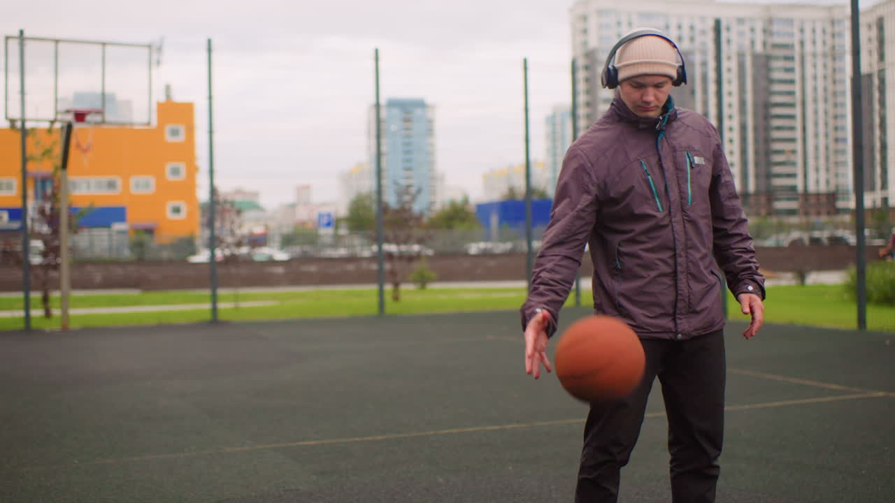Giovane uomo palleggia con un pallone da basket sul campo, indossa cuffie e un berretto, giacca viola, skyline della città sullo sfondo, cielo nuvoloso, espressione concentrata, si allena nel controllo della palla, allenamento urbano solitario