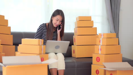 Una-Mujer-Joven-Que-Trabaja-En-Su-Computadora-Portátil-Está-Sentada-Entre-Montones-De-Cajas-Esperando-El-Envío-Mientras-Realiza-Múltiples-Tareas-Al-Contestar-Su-Teléfono-Celular