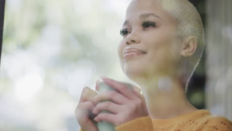 Happy-biracial-woman-drinking-coffee-at-window-at-home-in-slow-motion