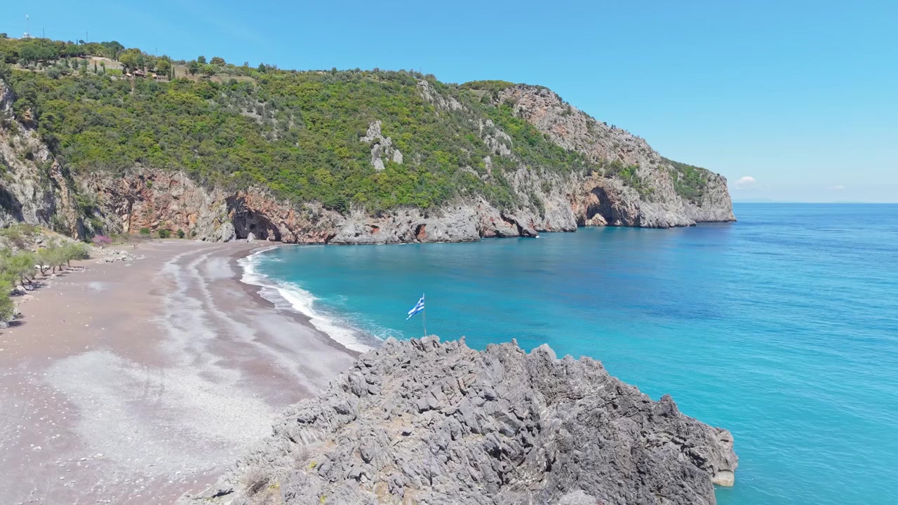 Riprese aeree con drone che circondano una bandiera greca mentre la mozzafiato spiaggia di Limnionas emerge sullo sfondo, mostrando acque turchesi e la costa frastagliata dell'Eubea