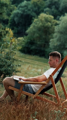 nice place to work remote in the nature. caucasian man works on his laptop sitting in a folding chair. side view. vertical video