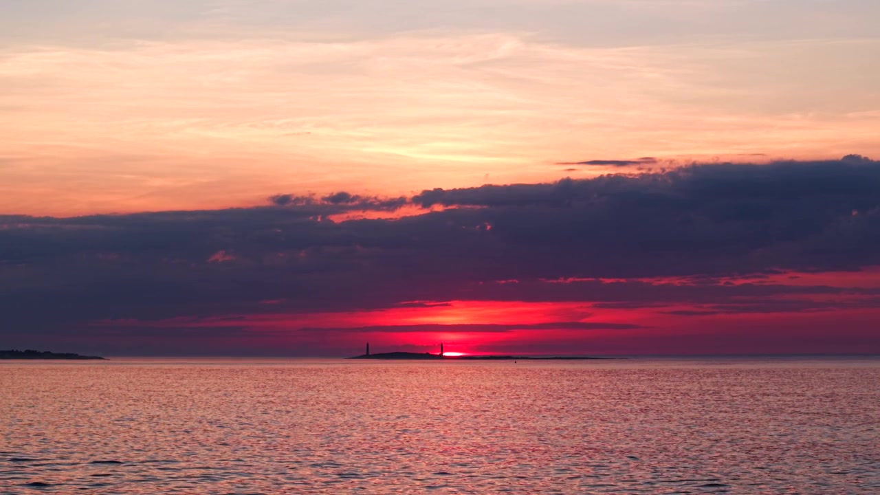 Sweeping wide shot of Thacher Island and twin lighthouses at sunset in Rockport, MA. Vibrant sky, glowing horizon, dramatic clouds, and gentle ocean waves create a serene, cinematic scene.