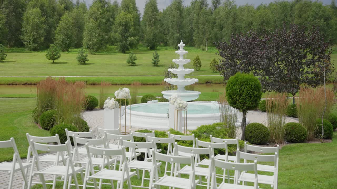 Beautiful outdoor wedding ceremony setup featuring white chairs and a decorative fountain, surrounded by lush greenery in a peaceful garden setting