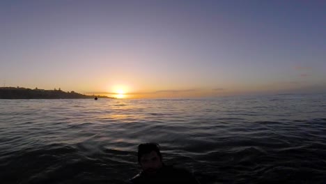 Young-man-surfer-sits-on-the-line-up-during-morning-surf-session,-waits-her-wave-and-enjoys-warm-tropical-sunrise