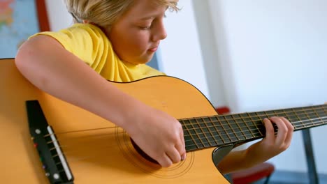 Blonder-Kaukasischer-Schuljunge-Sitzt-Am-Schreibtisch-Und-Spielt-Gitarre-In-Einem-Klassenzimmer-4k