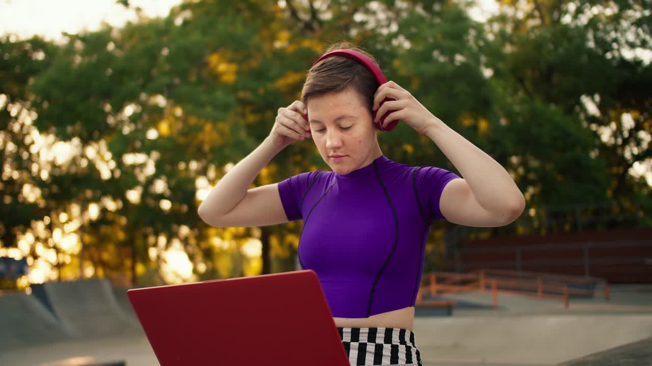 una joven de cabello corto en un top púrpura se pone auriculares rojos y comienza su trabajo con una computadora portátil roja en el parque en verano