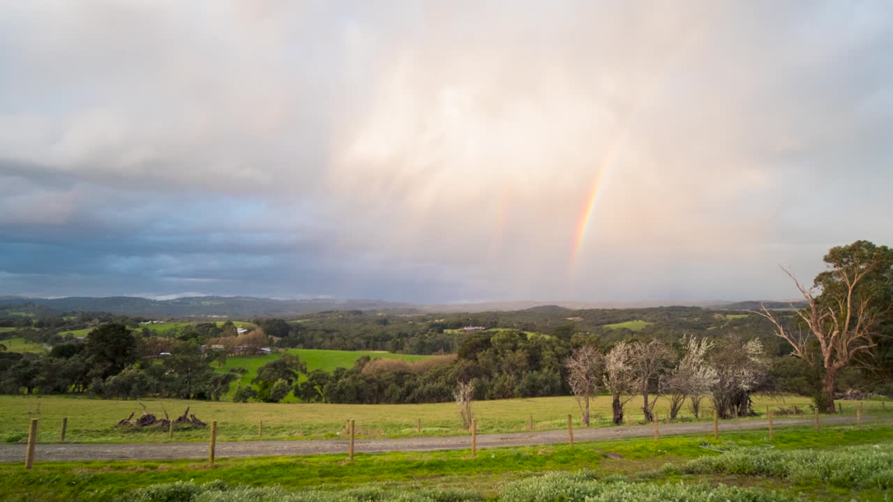 un hermoso lapso de tiempo de verdes colinas onduladas en australia con espesas nubes brillando a la luz del atardecer y un arco iris que aparece en el cielo