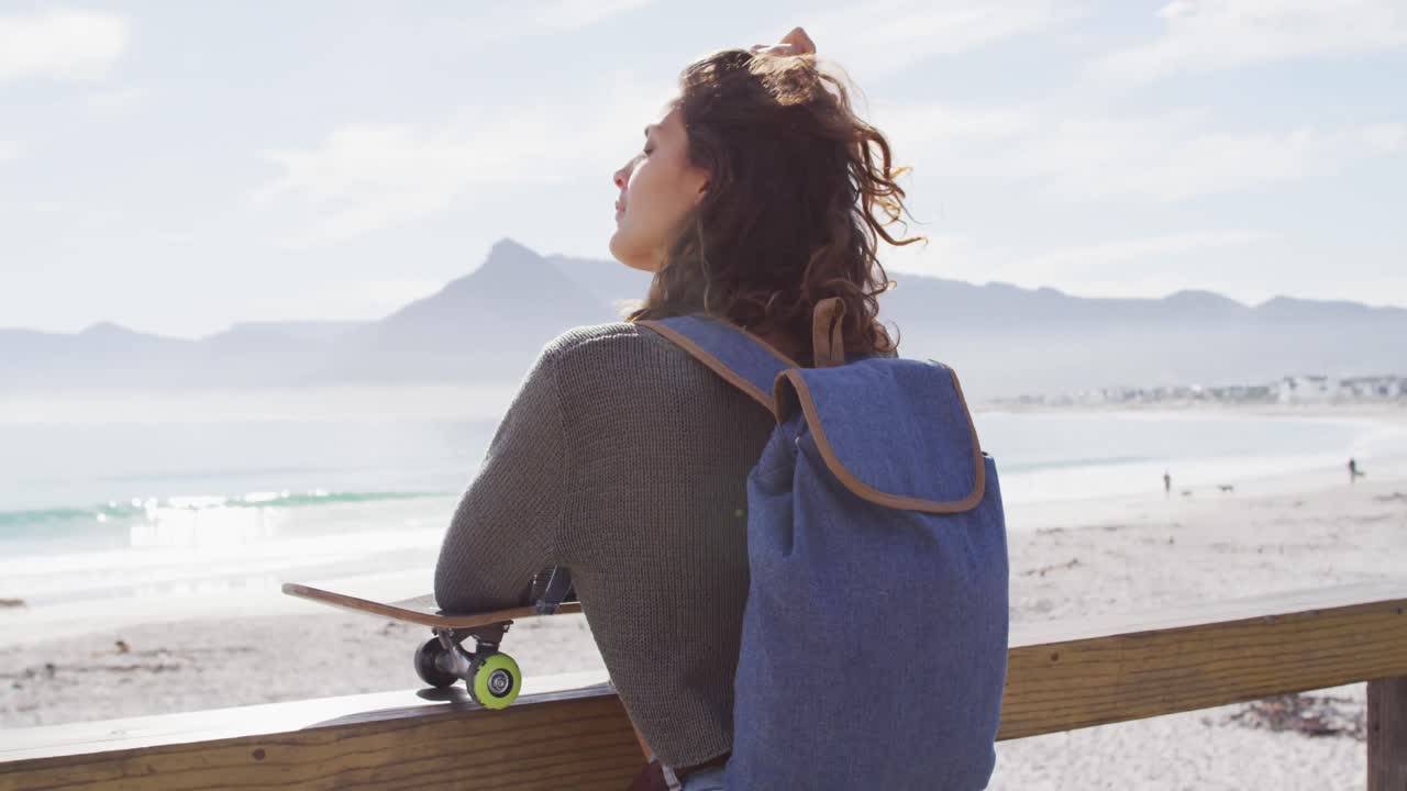 Donna multietnica che si appoggia a uno skateboard, in piedi al sole, mentre si gode la vista sul mare.