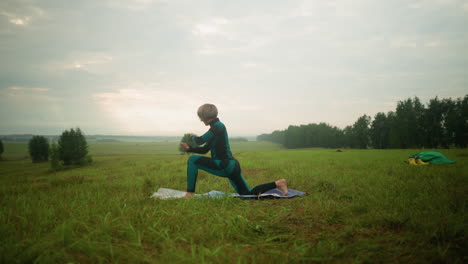 side view of lady in green and black suit outdoors in a vast grassy field practicing low lunge pose on a yoga mat, under cloudy sky with trees in the background and boots with green cloth nearby