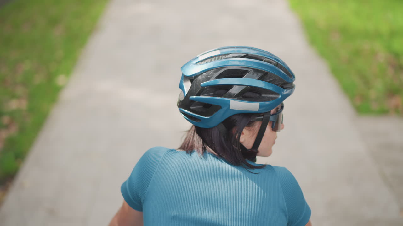 Signora con casco blu che pedala discretamente nelle vicinanze, donna serena in bici in un ambiente tranquillo, ciclista solitaria in abbigliamento blu che si muove silenziosamente lungo un marciapiede di periferia