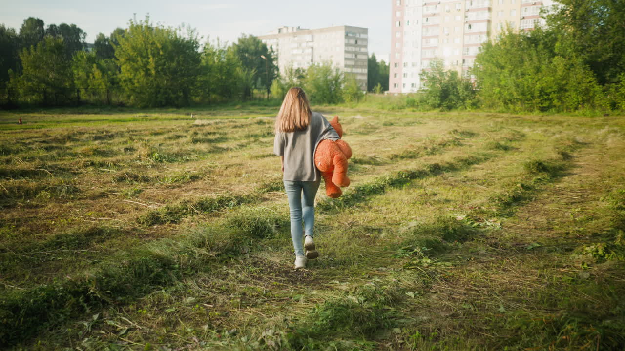 Rear view of young woman walking across open farmland carrying teddy bear under arm, surrounded by green vegetation and bordered by tall apartment buildings in distance under clear sky