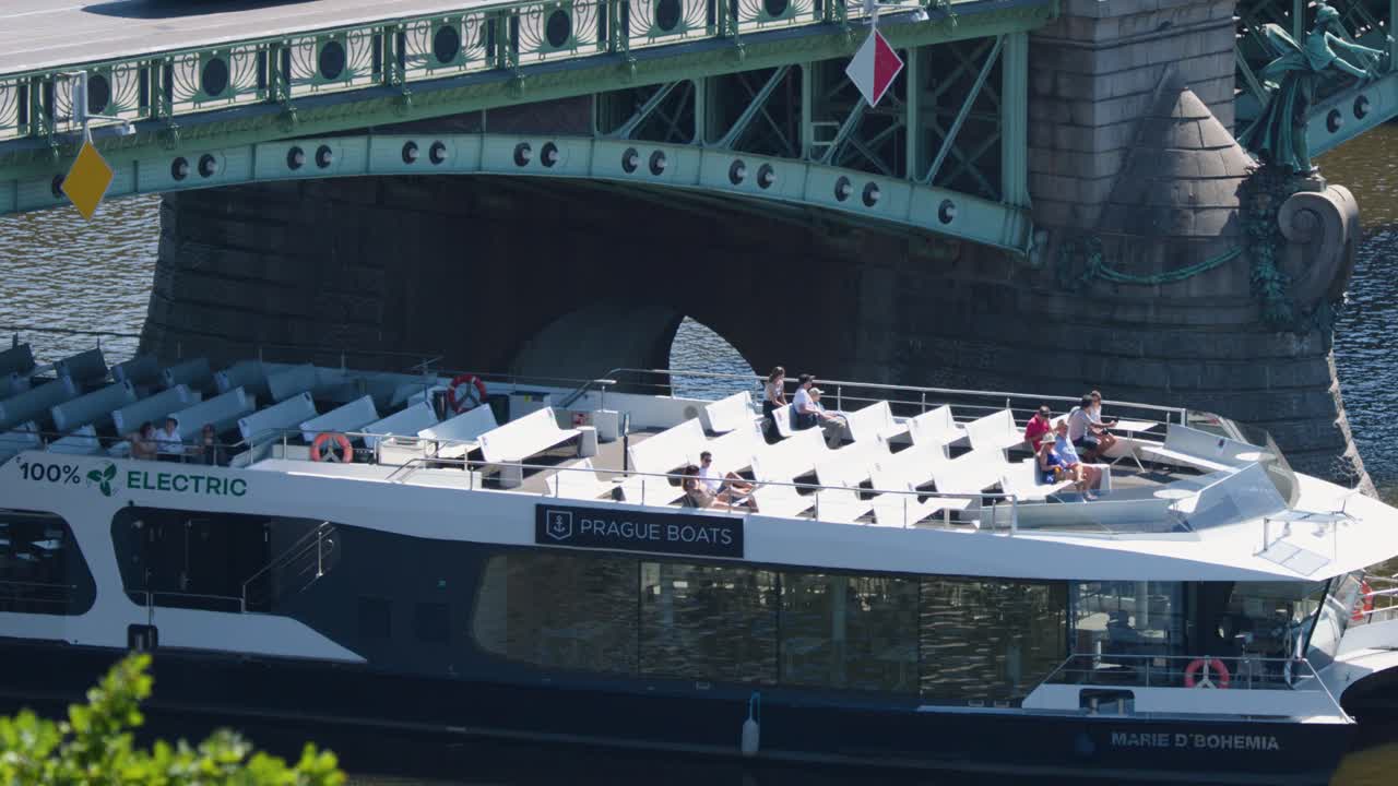 Modern electric boat with passengers cruises beneath historic bridge in Prague on sunny summer day