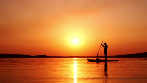 orange sunset over the river. silhouette of a male standing on the sup board and slowly rowing with a paddle.