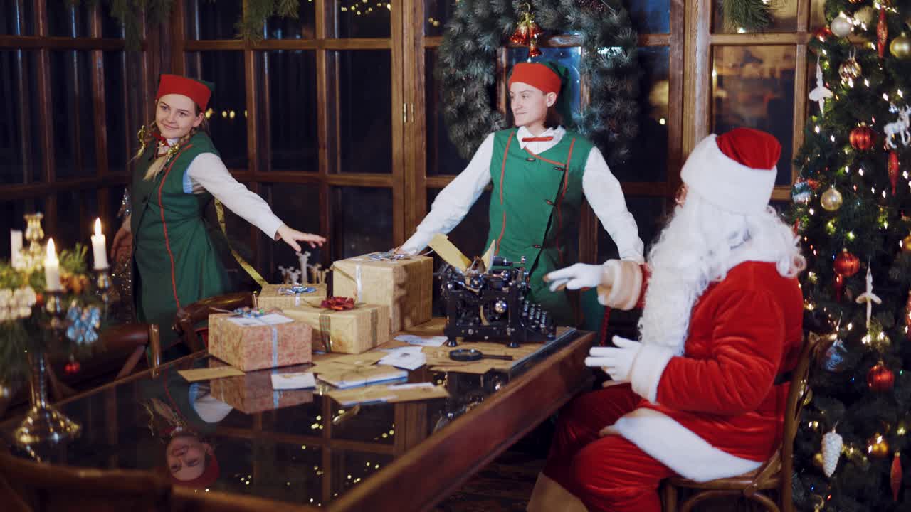Elves in festive costumes are dancing around the table with gifts, candles and a typewriter in the presence of Santa Claus, who watching and setting the rhythm on the background of the New Year tree near the window in the room.