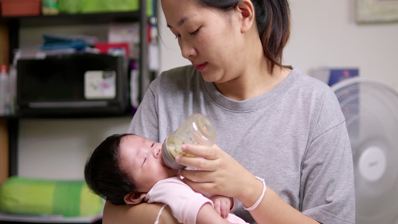 Sucking some milk from a baby bottle, a newborn child is falling in and out of sleep as its mother is holding her in her arms.