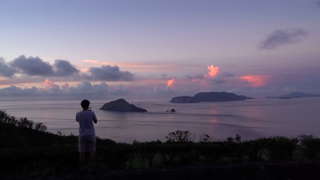 viajero masculino tomando fotos con su teléfono inteligente mirando hacia el océano abierto y las islas al amanecer con un hermoso resplandor de nubes