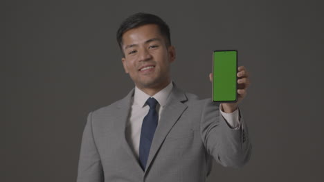 studio shot of smiling businessman in suit holding green screen mobile phone