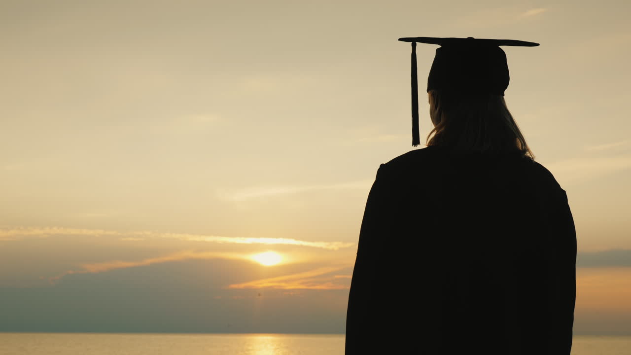un soltero con un diploma en la mano y una gorra de graduado mira el amanecer sobre el mar
