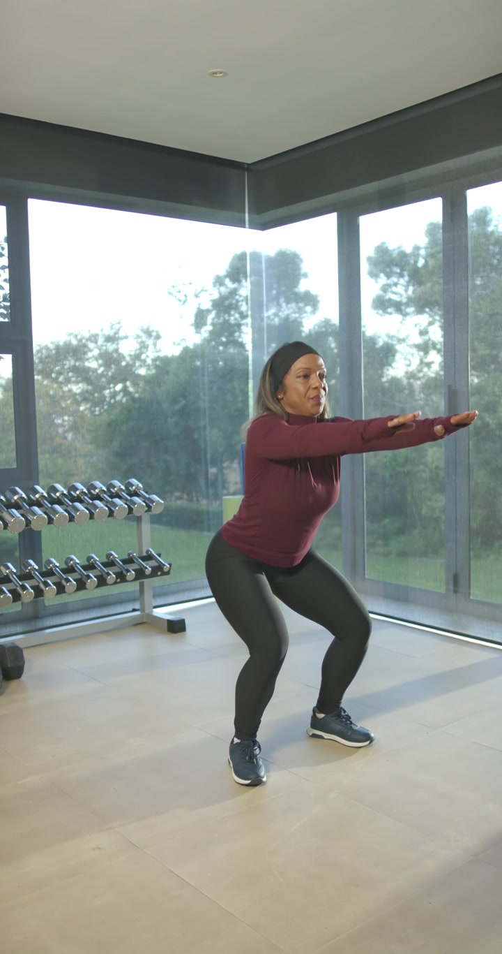 Video verticale: Donna afroamericana con top color bordeaux che fa squat per fitness in studio vicino ai manubri