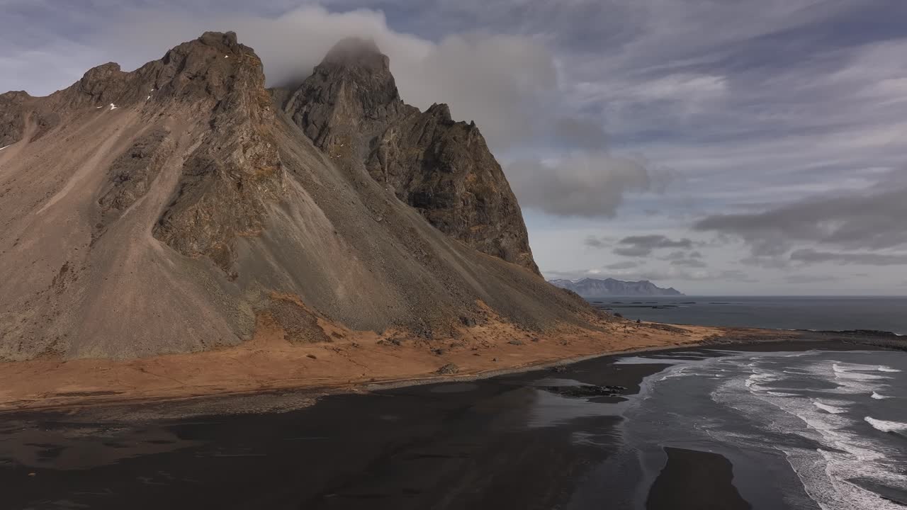 Drone pulling back to reveal the dramatic peaks of the Horny Mountains — Vestrahorn, Eystrahorn, and Brunnhorn — rising above the black sand beaches of Stokksnes, Iceland.